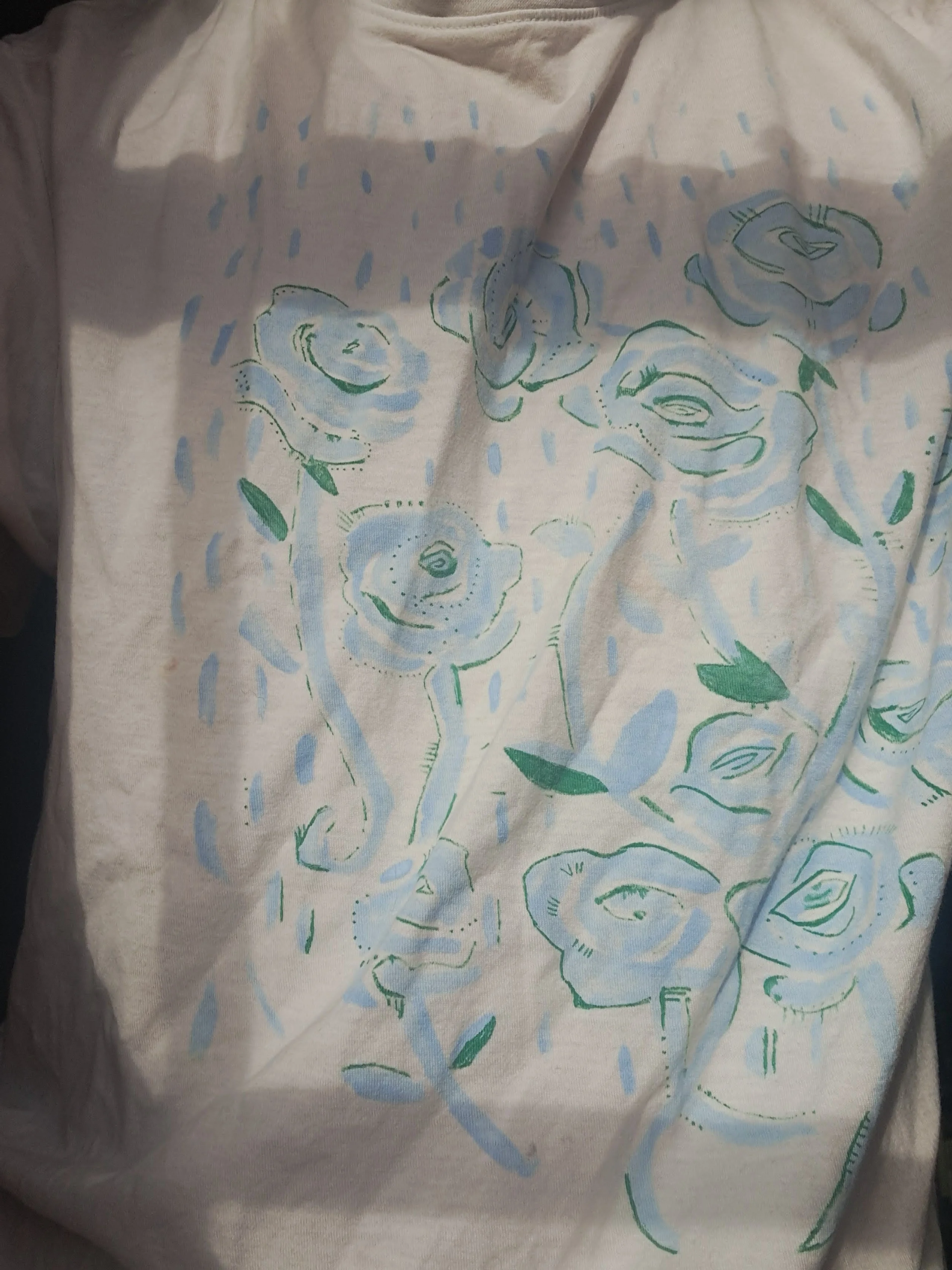 A white T-shirt painted with pale blue roses in a shower of rain. Details like the edges of the petals are dotted on top in green.